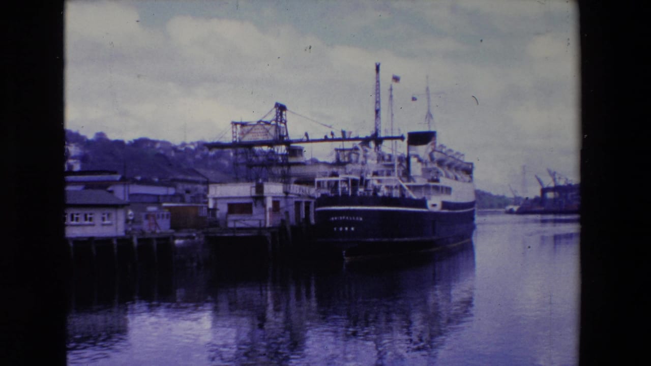 Ireland 1969 8mm frame of the INISFALLEN ferry at the pier, with a crane structure, shoreline buildings, calm water, and a cloudy sky visible around the vessel.