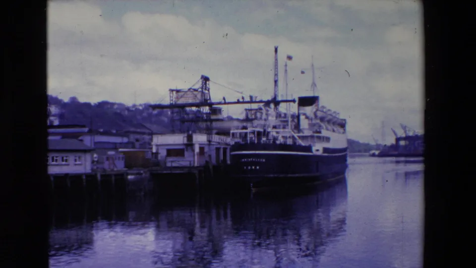 Ireland 1969 8mm frame of the INISFALLEN ferry at the pier, with a crane structure, shoreline buildings, calm water, and a cloudy sky visible around the vessel.