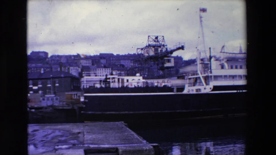 Ireland 1969 8mm frame showing a side view of the docked ferry, a foreground quay edge, industrial cranes, waterfront structures, and dense buildings on the hillside beyond.