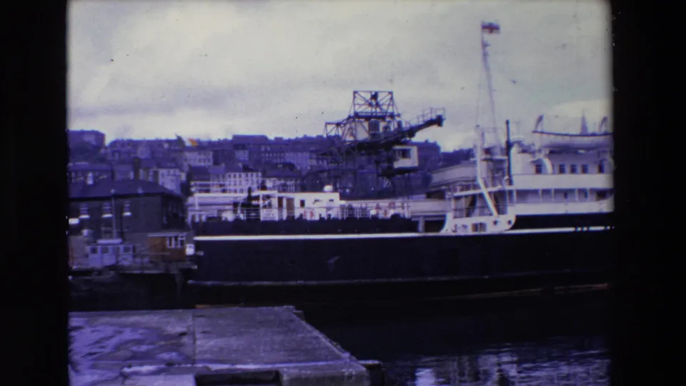 Ireland 1969 8mm frame of a large moored ferry beside the quay, with cranes rising behind the vessel and urban waterfront buildings visible under a pale overcast sky.