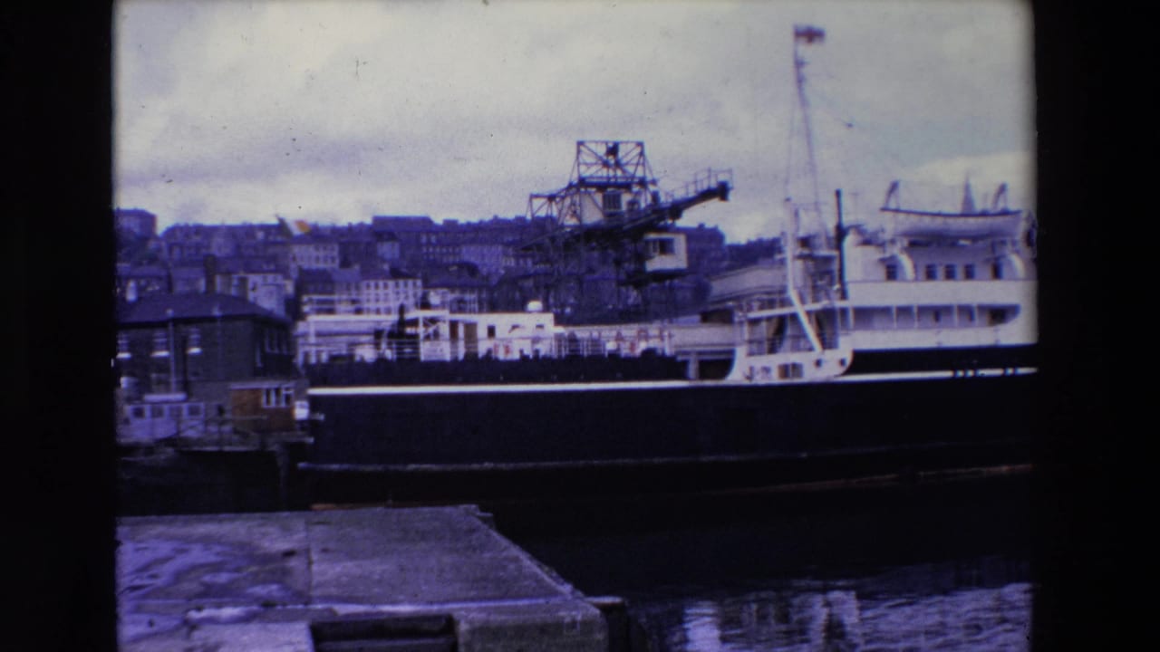 1969 Ireland home-movie frame showing a large docked ship, industrial waterfront crane, harbor water, quay edge, and hillside buildings under an overcast sky.