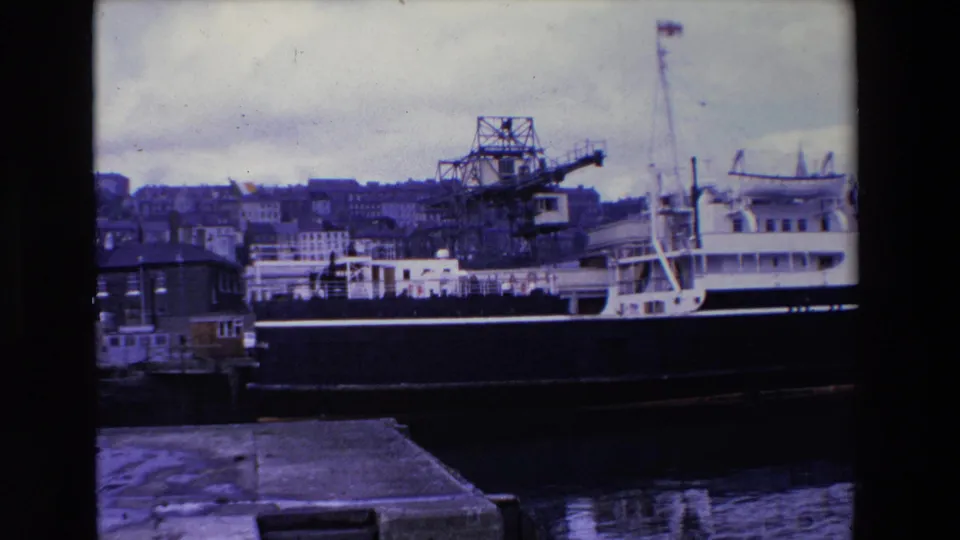 1969 Ireland home-movie frame showing a large docked ship, industrial waterfront crane, harbor water, quay edge, and hillside buildings under an overcast sky.