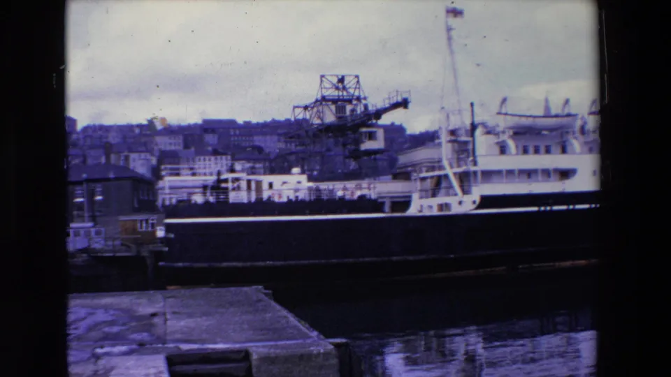 1969 Ireland frame with the ship held steady in view, showing the black hull, white upper deck, crane, harbor water, and buildings rising behind the port.