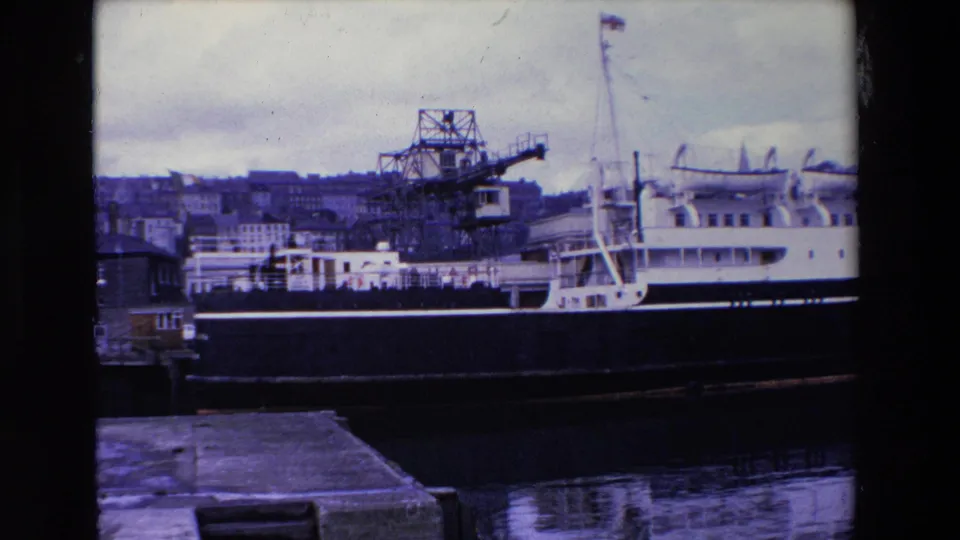 1969 Ireland home-movie still showing a docked cruiser-like vessel beside a crane, with a quay in the foreground and dense hillside buildings across the harbor.