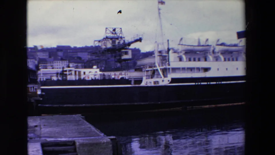 Final 1969 Ireland frame showing the docked ship and crane from a fixed home-movie viewpoint, with water reflections, quay stonework, and hillside port buildings visible.