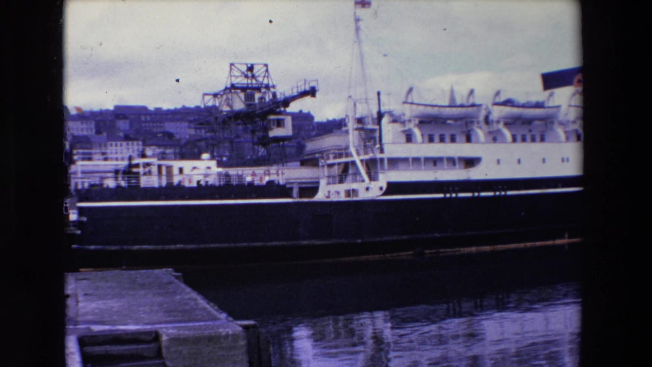 1969 Ireland 8mm frame showing a white multi-deck passenger vessel with a dark hull moored at a harbor, with industrial cranes and buildings in the background.