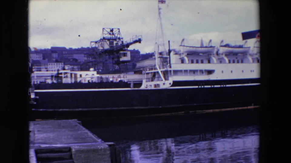 1969 Ireland 8mm frame showing a white multi-deck passenger vessel with a dark hull moored at a harbor, with industrial cranes and buildings in the background.