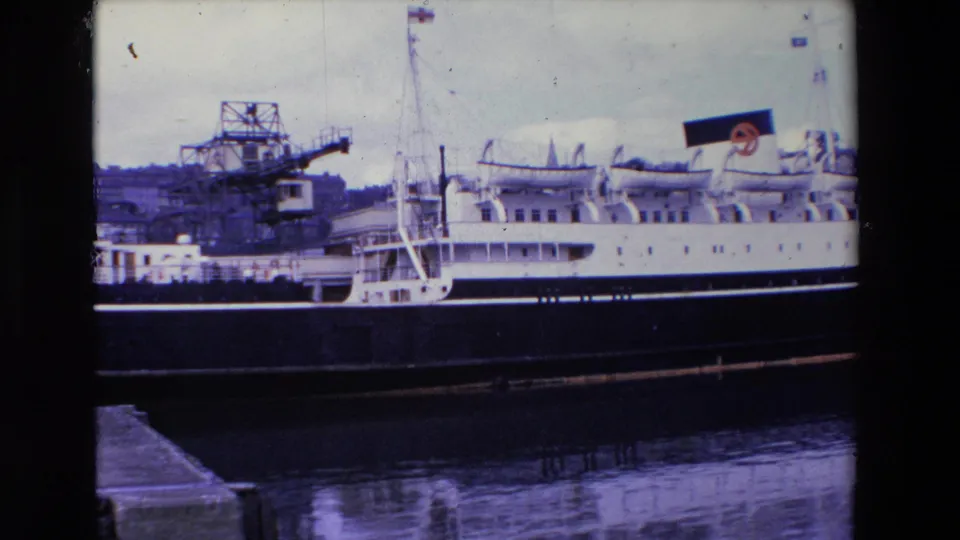 1969 Ireland 8mm frame showing the side of a docked white passenger ship, with lifeboats, a visible funnel marking, cranes, and calm water near the quay.