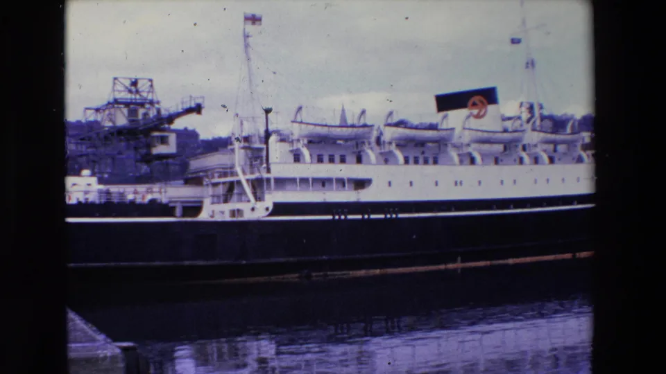 1969 Ireland 8mm frame of a moored passenger vessel with a dark hull and white upper decks, set against dockside cranes and reflected harbor water.
