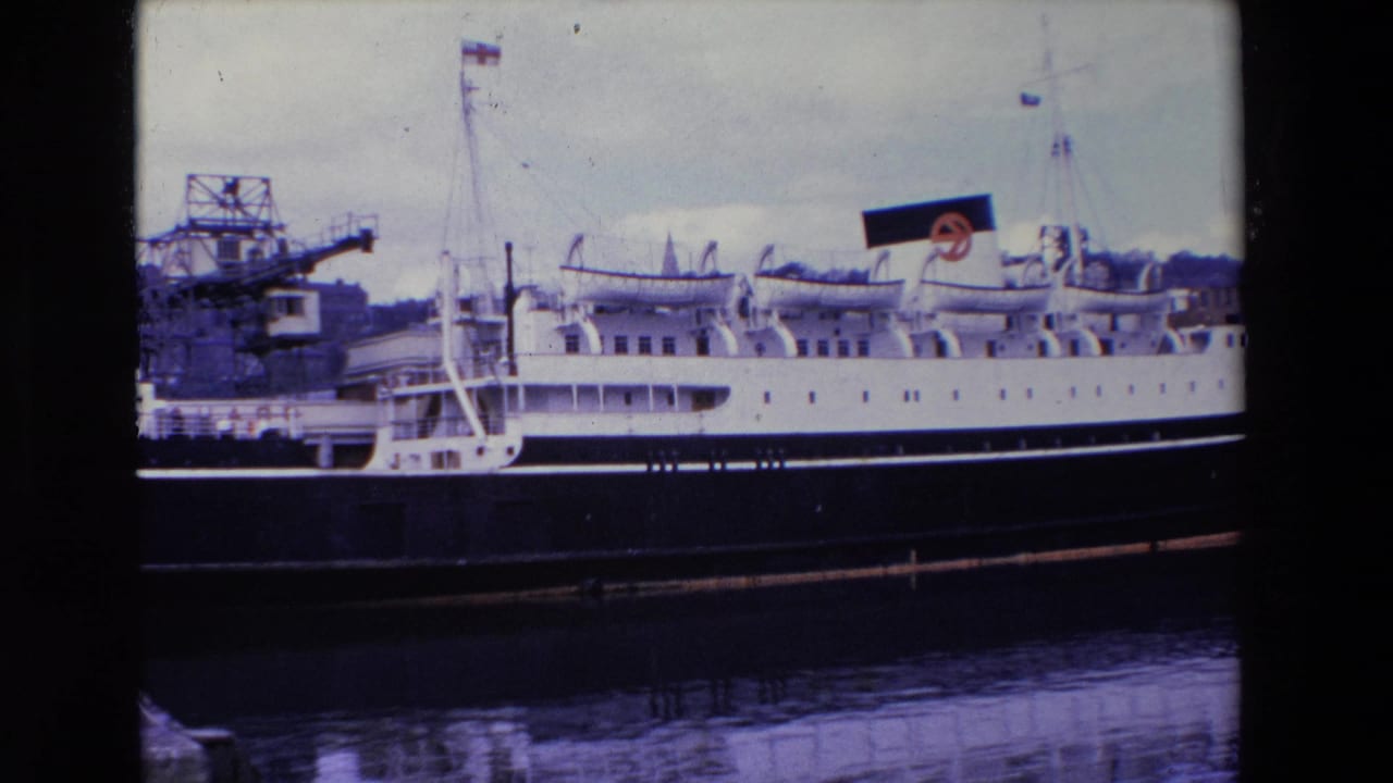 1969 Ireland 8mm frame showing a large white-and-black passenger ship moored beside a pier, with cranes, mast flags, and a reflected hull in the water.