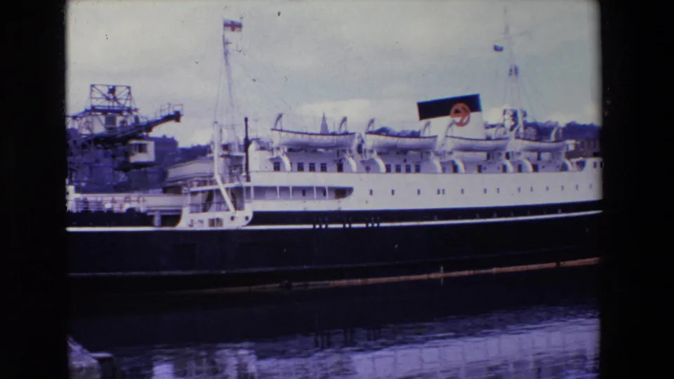 1969 Ireland 8mm frame showing a large white-and-black passenger ship moored beside a pier, with cranes, mast flags, and a reflected hull in the water.