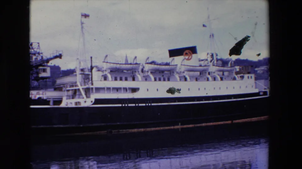 1969 Ireland 8mm frame with the passenger ship seen broadside at the pier, showing a marked funnel, industrial harbor structures, and calm reflective water below.