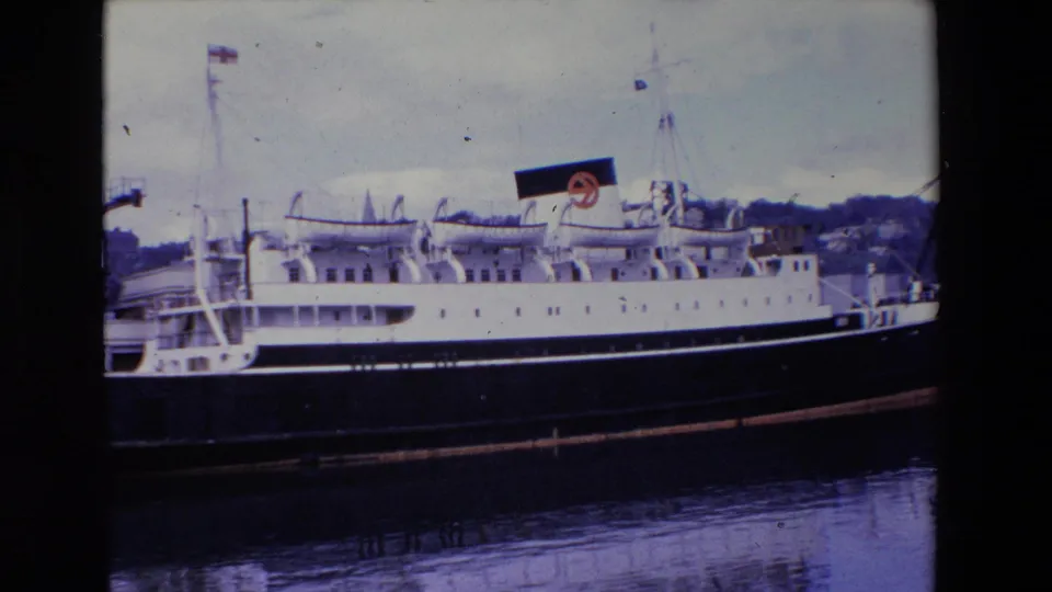 1969 Ireland 8mm frame showing the same moored passenger ship, with its dark hull mirrored in the water and a low shoreline visible beyond the pier.