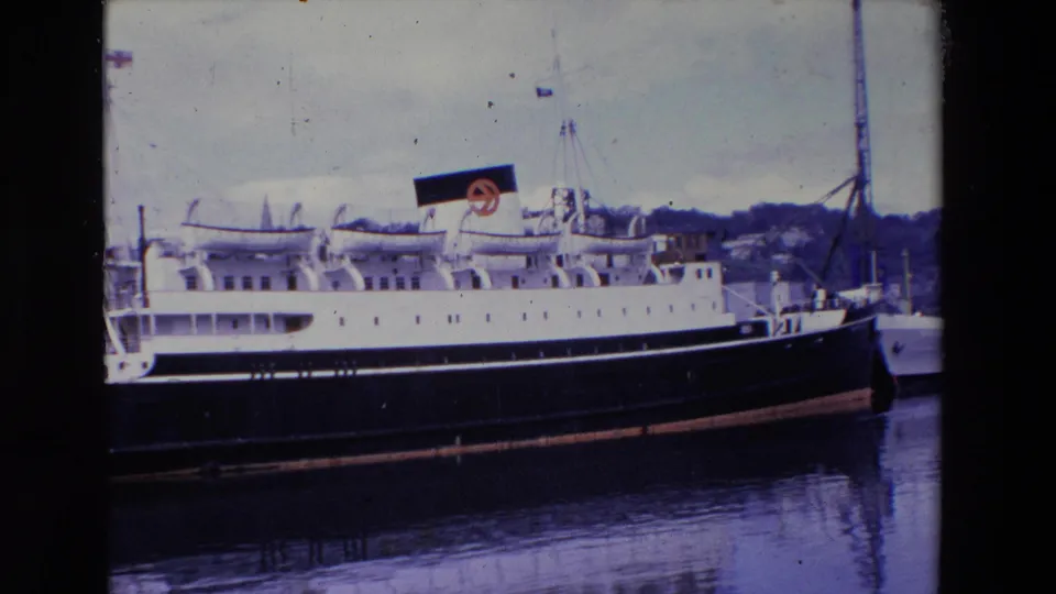1969 Ireland 8mm frame showing the ship near the end of the shot, with its bow area, funnel marking, harbor mast, pier, and dark reflection visible in the water.