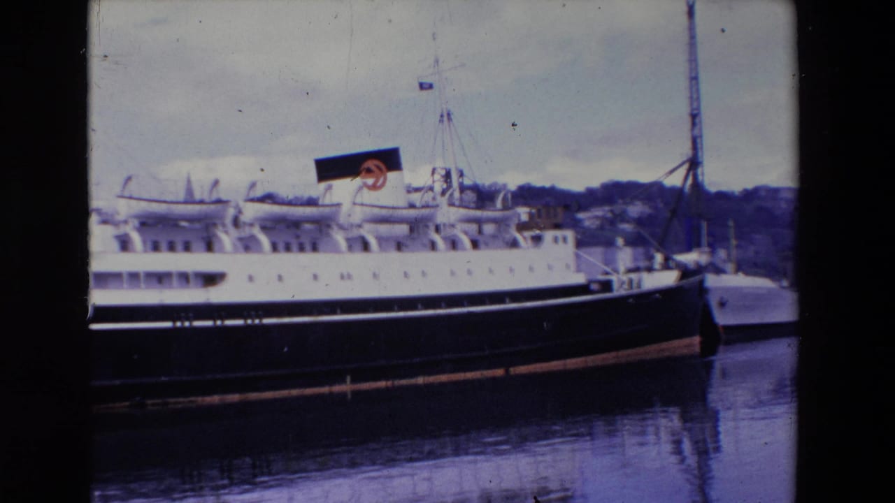 1969 Ireland 8mm frame showing a large passenger liner at dock, with a black funnel bearing a red circular logo, calm water, and a hilly shoreline in the background.