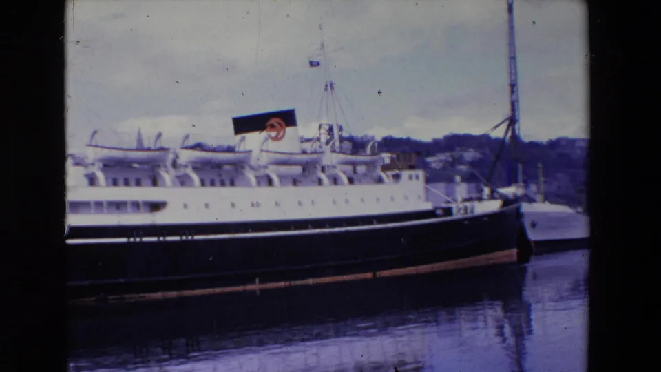 1969 Ireland 8mm frame showing a large passenger liner at dock, with a black funnel bearing a red circular logo, calm water, and a hilly shoreline in the background.