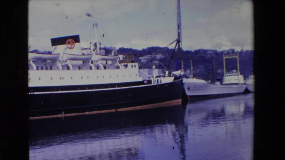 1969 Ireland 8mm frame showing the liner closer to the bow area, with a smaller vessel alongside, a tall crane, and hillside buildings beyond the harbor.