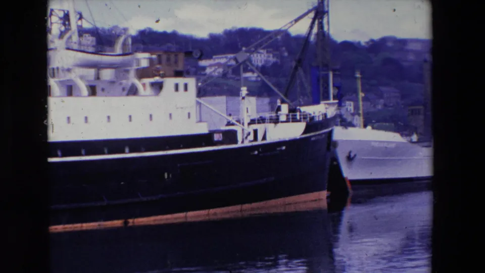 1969 Ireland frame from home-movie footage showing a closer harbor view of the liner's forward section, a moored smaller vessel, dock crane, and hillside beyond.