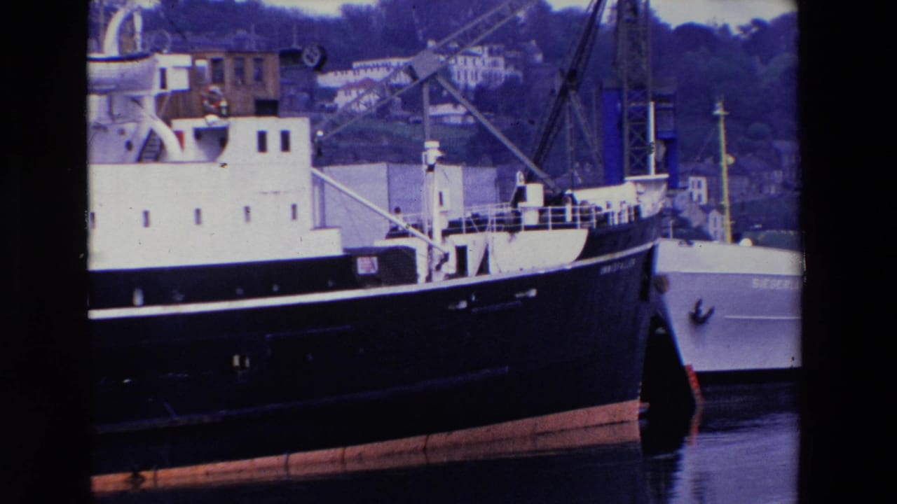 1969 Ireland 8mm frame showing a large docked ship with a white superstructure, dark hull, port cranes, and hillside buildings in the background.