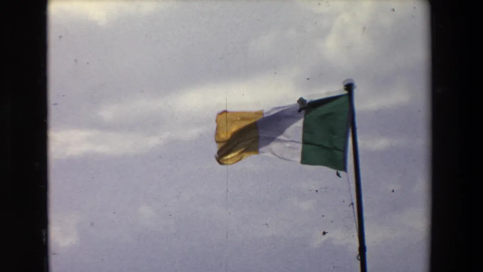 Irish tricolor flag on a pole waving in wind against a cloudy blue-gray sky in 1969 Ireland home-movie footage.