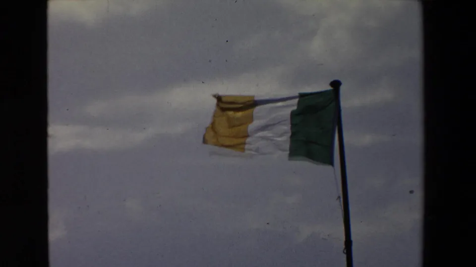 The Irish tricolor continues to flap on a pole under overcast blue-gray sky, with grain and soft focus from the 8mm source.