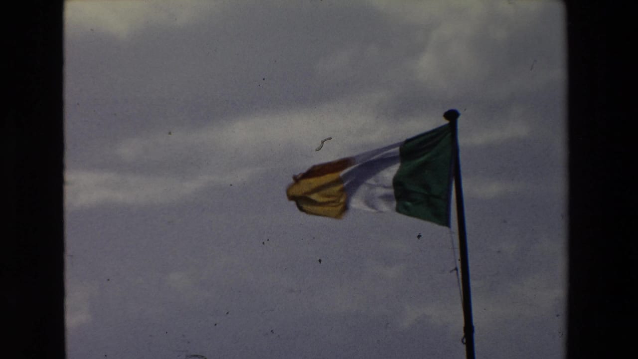 Irish tricolor flag waves in the wind against a cloudy sky in 1969 Ireland home-movie footage, with 8mm frame borders visible.