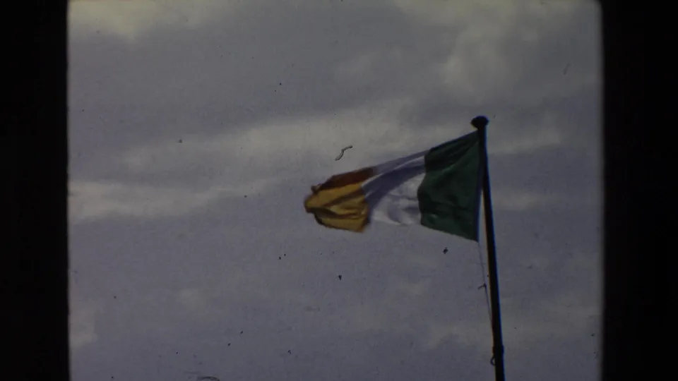 Irish tricolor flag waves in the wind against a cloudy sky in 1969 Ireland home-movie footage, with 8mm frame borders visible.