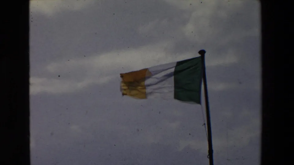 Wind pulls the Irish tricolor outward from a flagpole against a gray-blue sky, with visible 8mm grain and edge borders.
