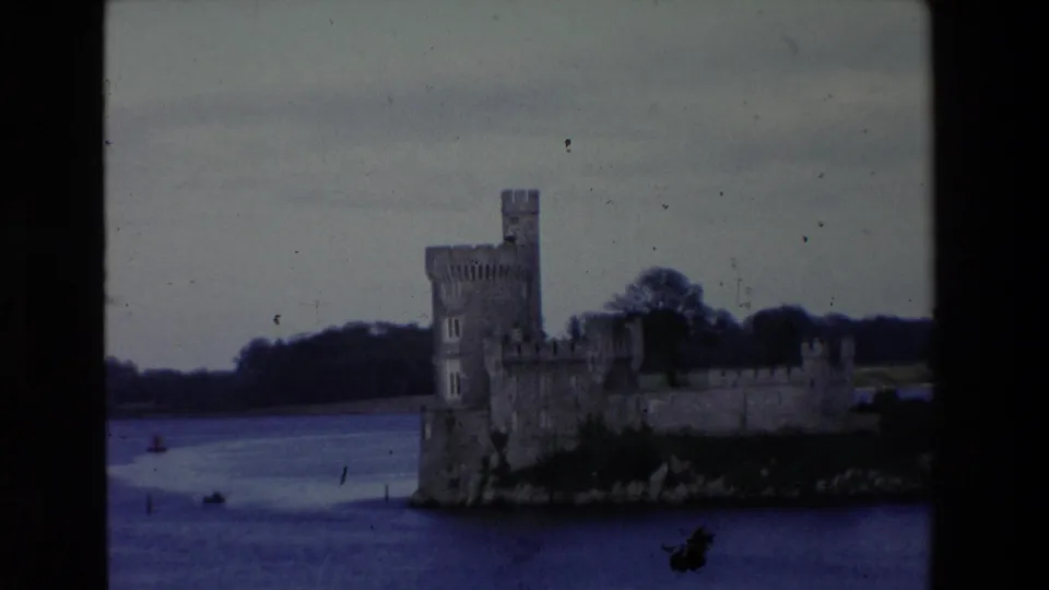 A stone castle and tower stand on a rocky lakeside outcrop in Ireland, with water, distant trees, and a small boat or buoy visible nearby.
