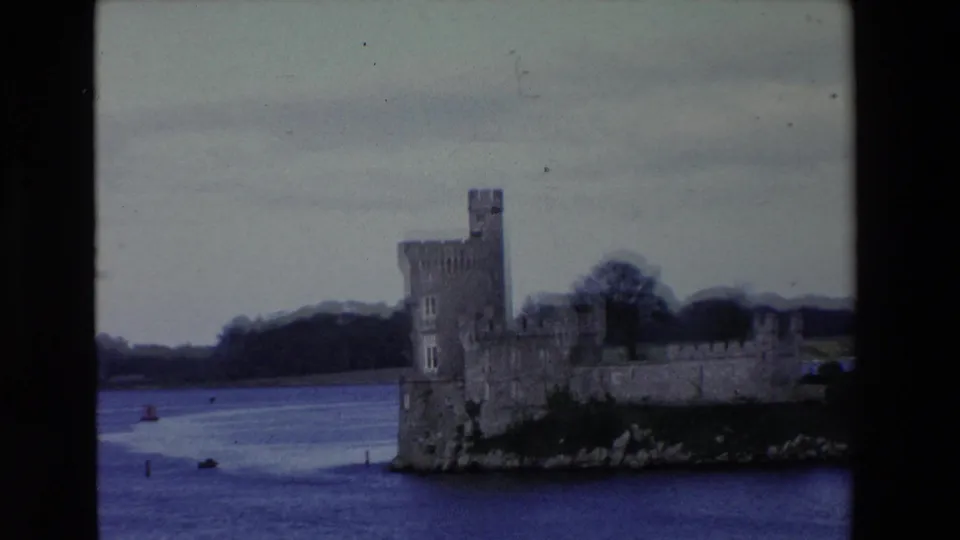 The castle remains framed across the lake, showing crenellated stone walls, a tower, shoreline rocks, and muted 8mm color from the 1969 Ireland reel.