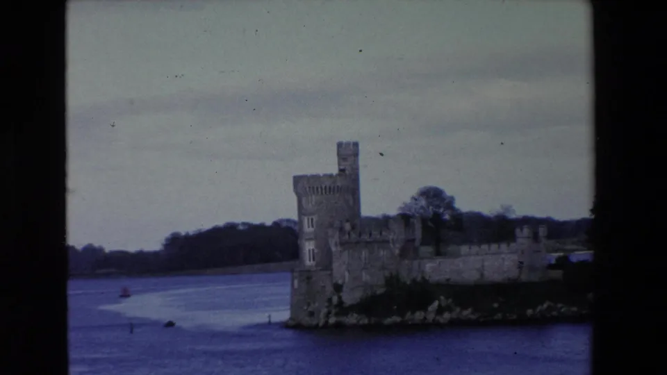 1969 Ireland 8mm frame showing the castle’s tower and long stone wall receding along the river, with wooded land behind it.