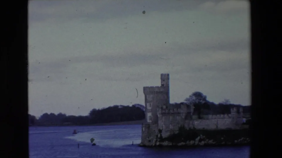 1969 Ireland 8mm frame with the castle partly right of center, broad river water in front, and a wooded shoreline under a pale overcast sky.