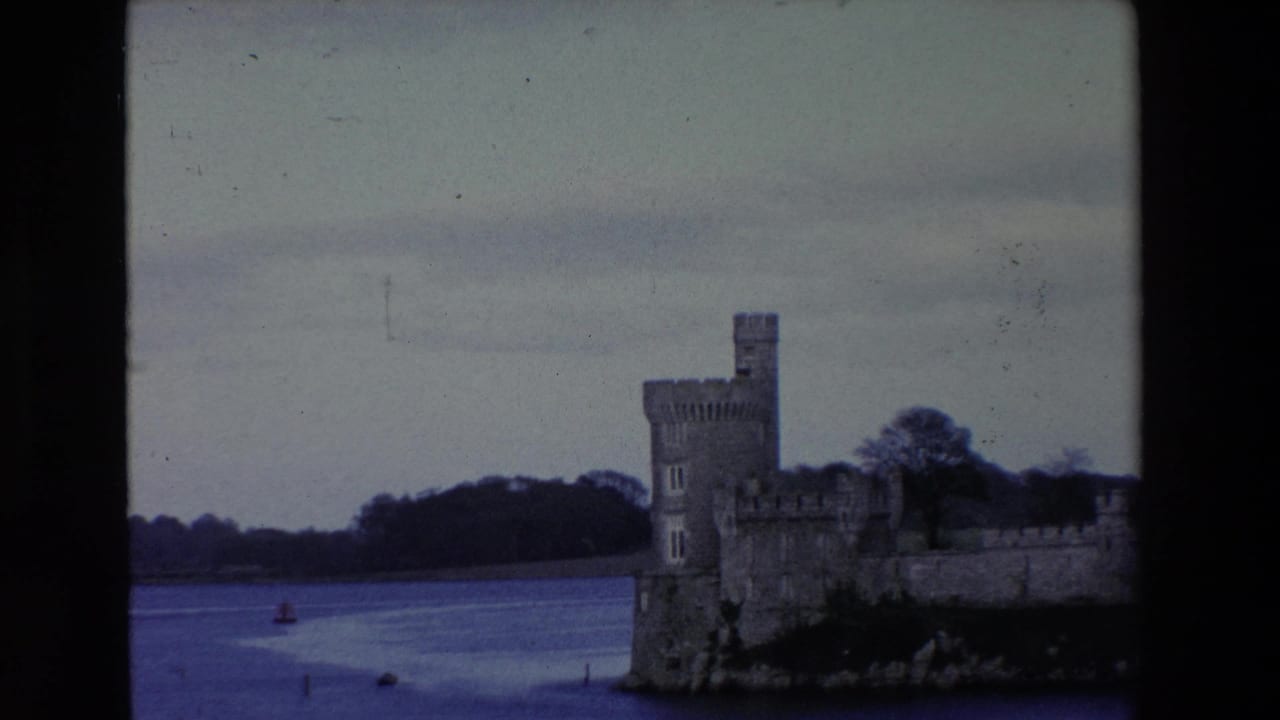 1969 Ireland 8mm frame showing a stone castle tower and adjoining wall beside calm water, with wooded shoreline and a small red buoy visible.