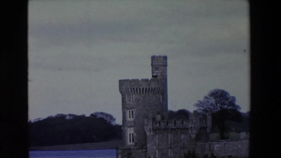 1969 Ireland home-movie frame with a closer view of the crenellated stone tower, castle wall, waterline, trees, and overcast sky.