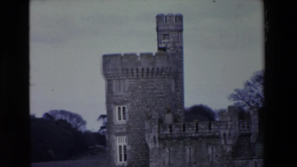 1969 Ireland 8mm frame focused on the castle tower, showing masonry, narrow windows, crenellations, adjoining wall, and trees in soft blue-purple color.