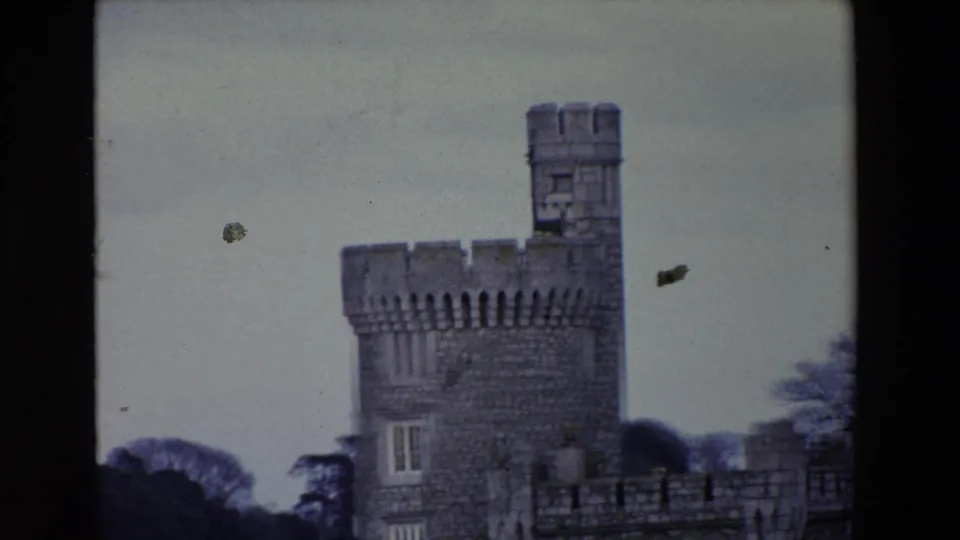 1969 Ireland home-movie frame showing the upper portion of the stone tower and wall against a cloudy sky, with visible dust marks from the film scan.