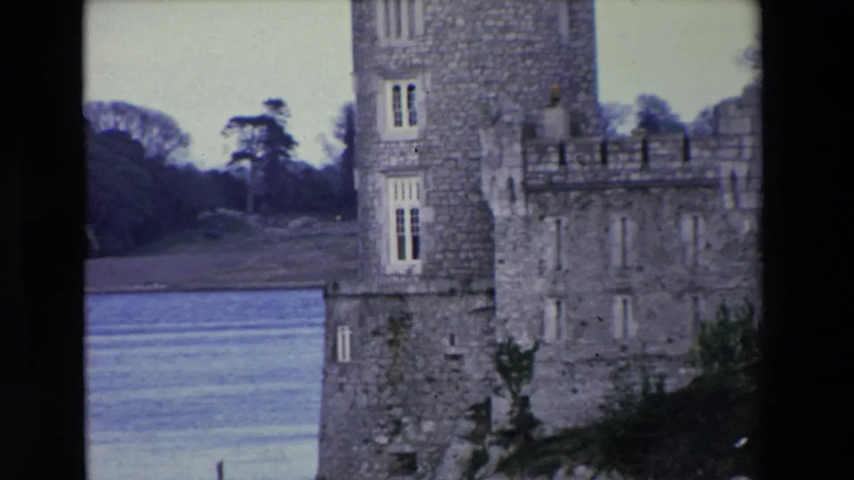 1969 Ireland 8mm frame with the camera closer on the stone castle facade, showing tall windows, battlements, river water, and a tree-lined background.