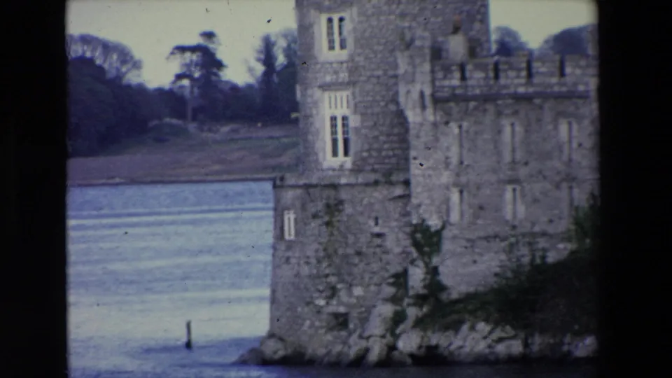 1969 Ireland home-movie frame showing the castle tower and adjoining wall beside broad calm water, with rocks or shoreline at the base and trees behind.