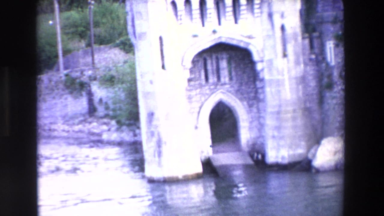 1969 Irish home-movie frame of a stone castle entrance at the lakeside, with a pointed arch, short ramp, shoreline rocks, and reflective water below.