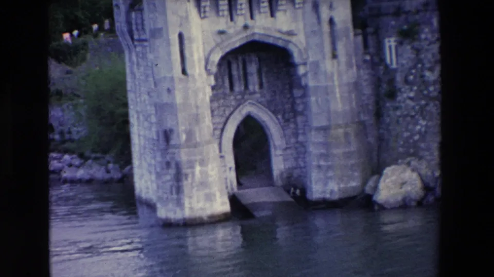 Closer 1969 Irish home-movie view of the lakeside castle entrance, showing the pointed arch, stone supports, ramp, water surface, and rocks near the wall.