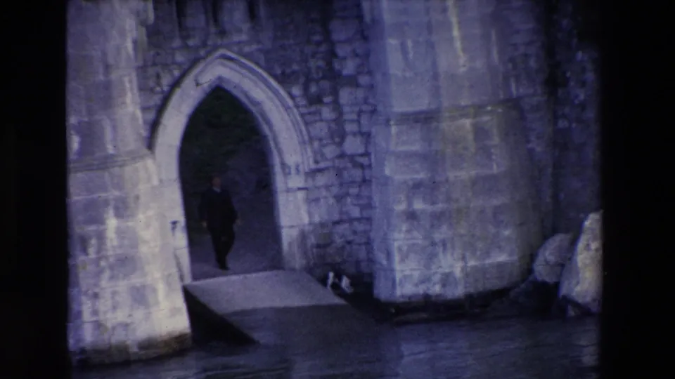 1969 Irish home-movie frame showing an adult man briefly visible inside the castle's arched entrance, with the ramp, stone facade, rocks, and water in the foreground.