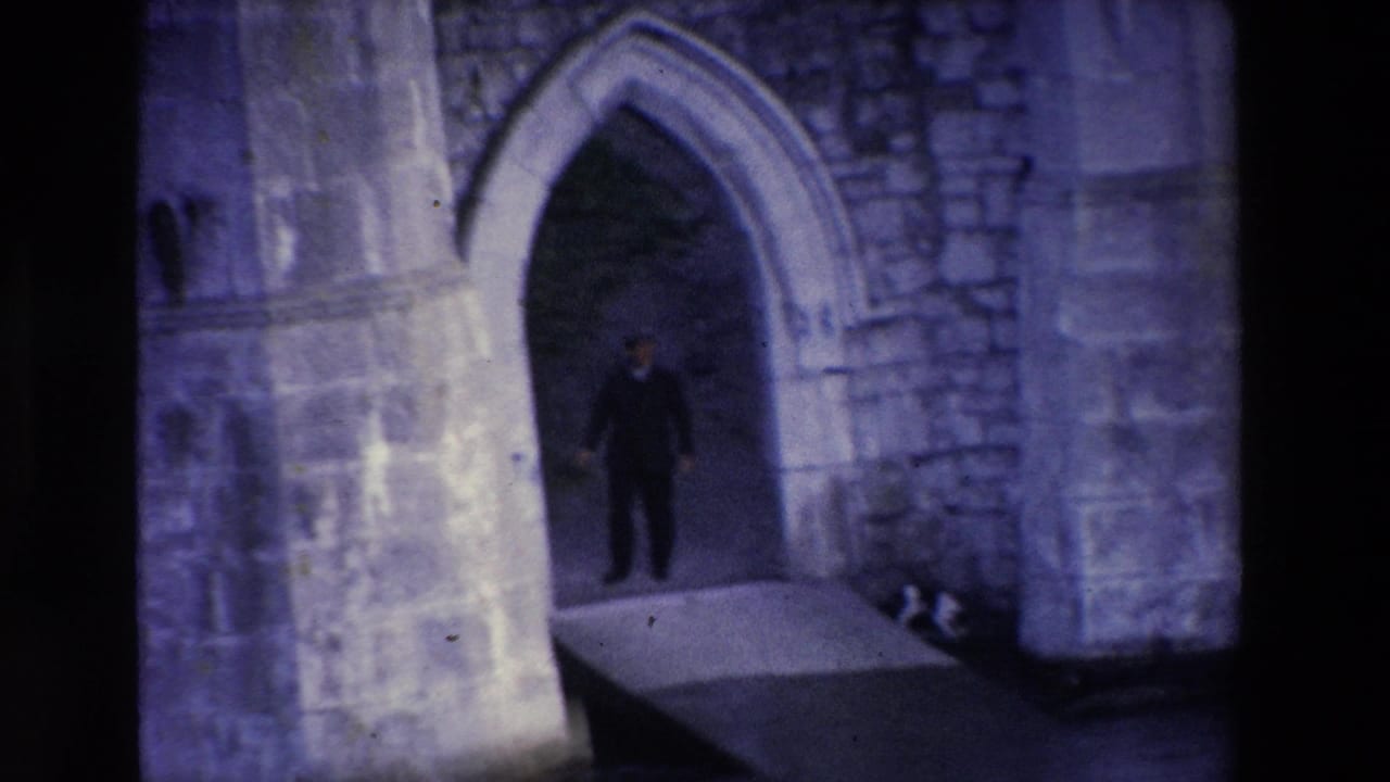 In 1969 Ireland home-movie footage, a man in a dark suit stands in shadow within a Gothic stone archway beside rough masonry walls.