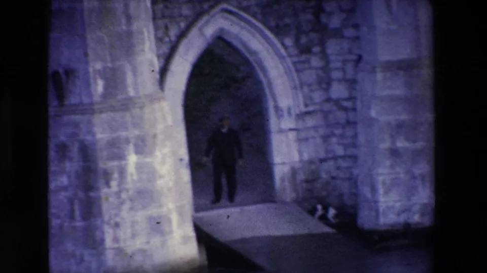 In 1969 Ireland home-movie footage, a man in a dark suit stands in shadow within a Gothic stone archway beside rough masonry walls.