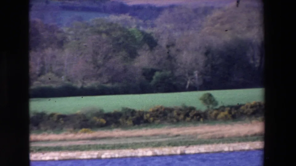 Ireland 1969 8mm frame showing blue water below a pale bank, with a hedgerow, green field, trees, and hillside in the background.