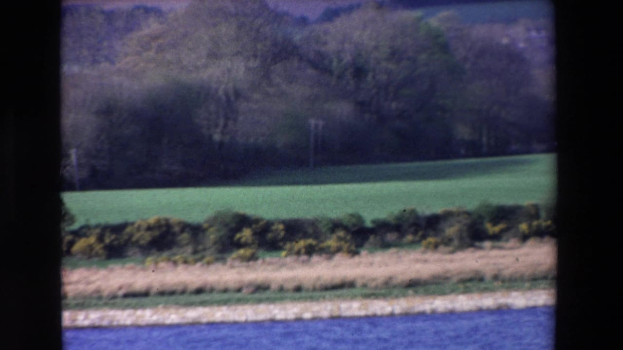 1969 Ireland 8mm frame showing a blue river in the foreground, hedgerows and green fields beyond, viewed through dark vertical window framing.