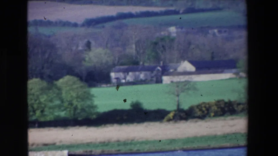 1969 Ireland 8mm frame showing distant farm buildings across green fields, with hedgerows, bare trees, and a blue river at the lower edge.
