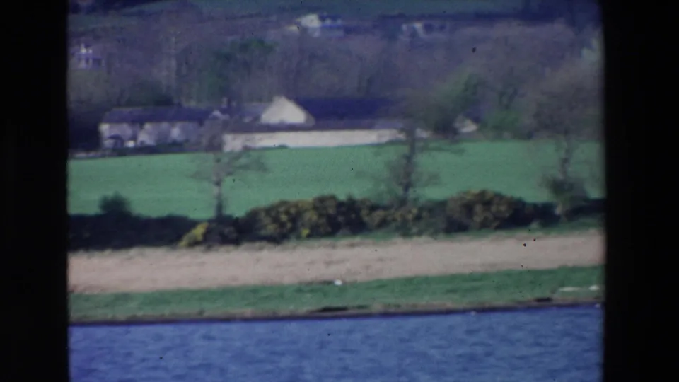1969 Ireland countryside frame with the river in the foreground, a pale field strip, hedgerows, green pasture, and distant farm buildings under wooded hills.