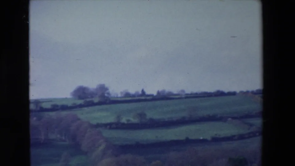 1969 Ireland 8mm frame of rolling meadow fields divided by hedgerows or stone walls, with distant trees under a soft grey sky.