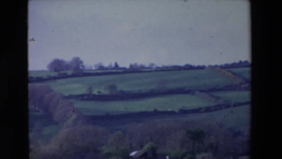1969 Ireland 8mm frame showing sloped green meadows, field divisions, distant tree lines, and soft home-movie blur from an elevated countryside viewpoint.