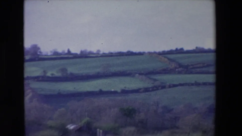 1969 Ireland 8mm frame of layered green fields and hedgerow or stone-wall boundaries, with a pale sky and soft film grain visible across the scan.